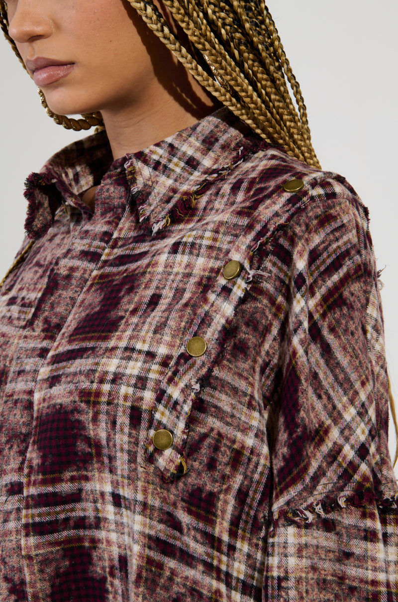 An individual is dressed in the DRIFTER FLANNEL, an oversized grunge shirt characterized by its brass buttons and frayed edges. This flannel showcases a blend of burgundy, cream, and brown tones with distressed detailing. The person has long braided hair and a neutral expression.
