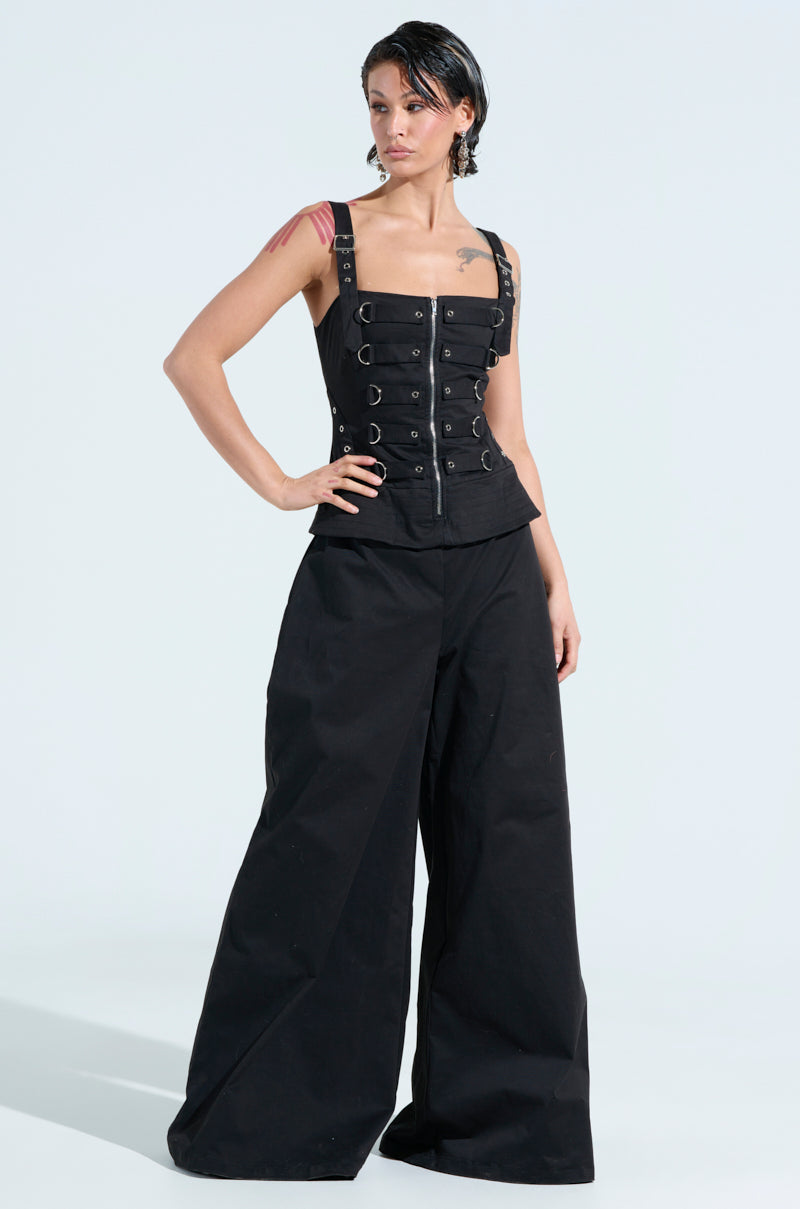 A person exudes confidence in an AMELIA JUMPSUIT WITH METAL DETAILS, featuring a black corset style and wide-leg pants. Their wet short hair and arm tattoos stand out against the plain white background.