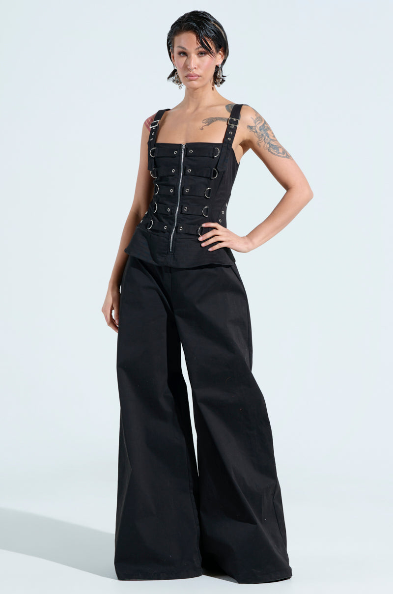 Sporting the AMELIA JUMPSUIT WITH METAL DETAILS, a person with short hair and visible tattoos stands confidently. The stylish ensemble with wide-leg trousers and metal accents is strikingly highlighted against a plain background, showcasing bold fashion choices.