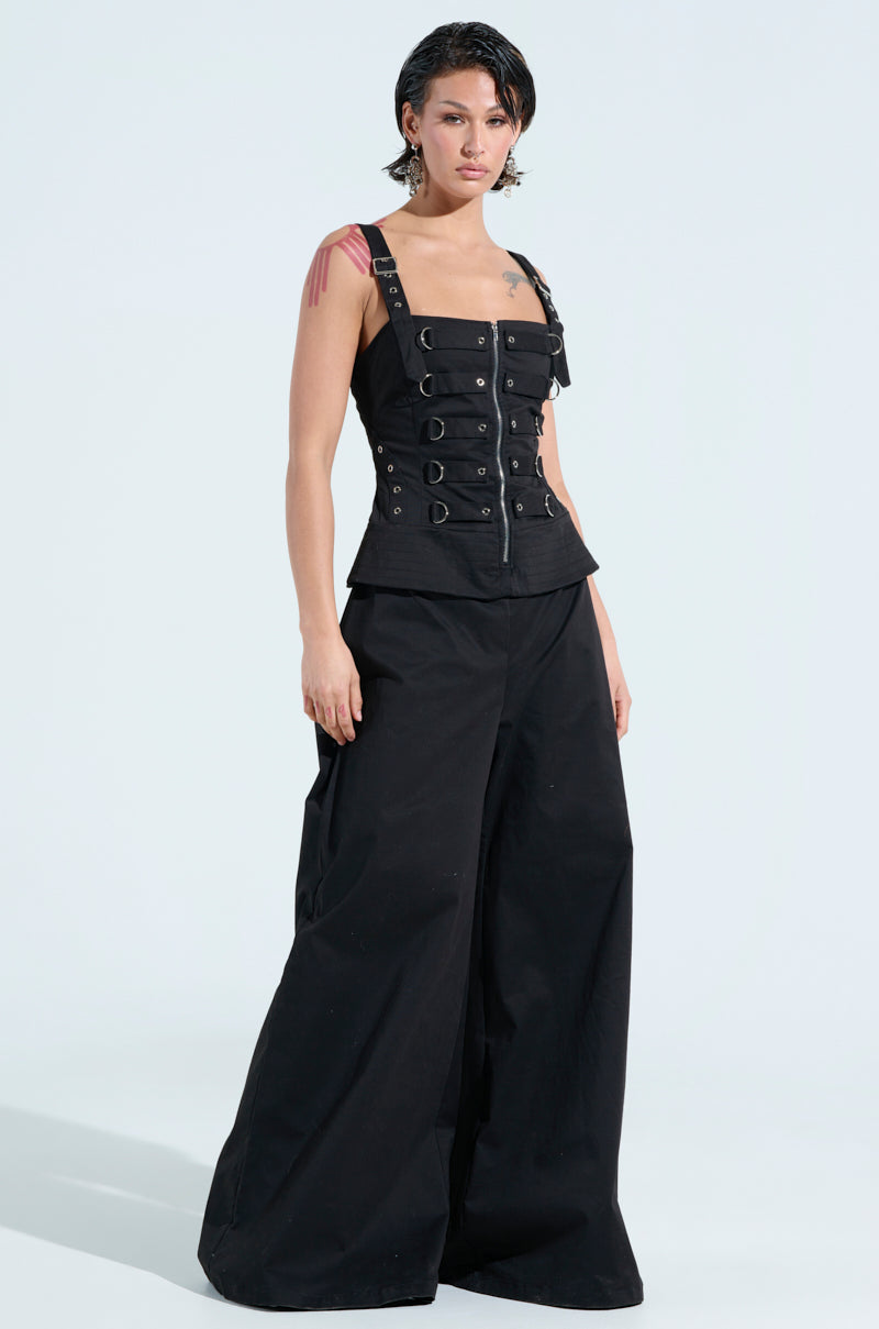 Dressed in the AMELIA JUMPSUIT WITH METAL DETAILS, a person with short, slicked-back hair confidently poses against a plain, light background.