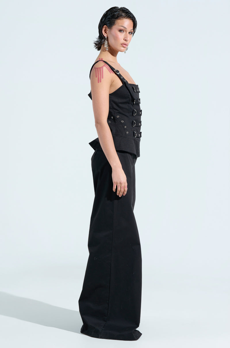 Against a light background, a person in profile sports the Amelia Jumpsuit with metal details. The outfit includes a black sleeveless top and wide-leg pants. They have short, dark hair and an upper arm tattoo.