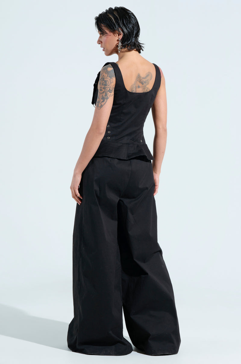 A person with short black hair, showcasing tattoos on their arm, stands in an Amelia Jumpsuit with Metal Details. They face away, highlighting its sleek sleeveless top and wide-leg pants against a light background.