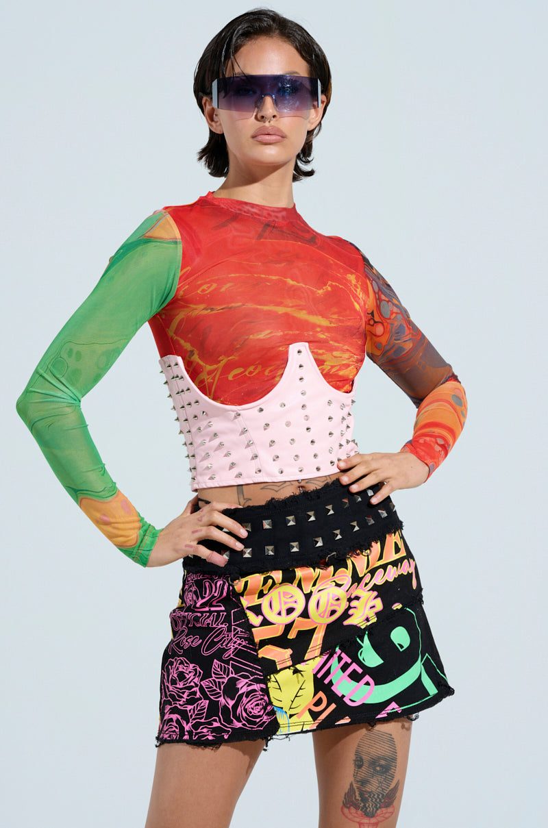 A person in a vibrant, patterned long-sleeved top, a pink studded corset, and a colorful graffiti-style skirt completes their look with faux leather sunglasses and the PRETTY BUT I BITE SPIKE BELT, standing confidently against a light background. 2