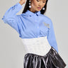 A woman in a blue striped shirt with oversized pearl earrings poses with one hand on her head. She is wearing the PAIR ME CORSET BELT in white over a metallic silver skirt, which features a lace-up back. Her gaze is downward against a plain background.