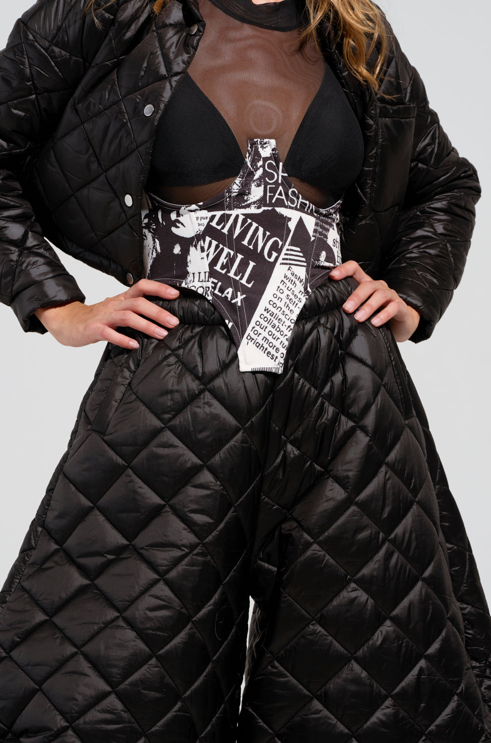 A person wears a black quilted jacket and pants ensemble, paired with a semi-transparent mesh top. The outfit includes a black bralette and is accentuated by the SPREAD THE NEWS SATIN UNDERBUST BELT, which features graphic white text and a wired underbust design. Standing with hands on hips, they showcase their distinctive fashion style.