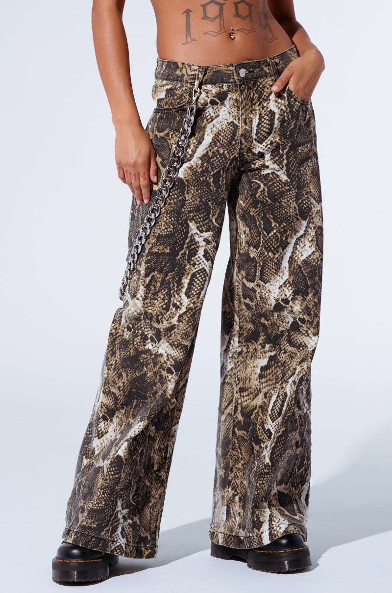 The person is wearing RUN AWAY SNAKE PANT with a high-rise waist and brown snakeskin design, complemented by a silver chain accessory. They are standing on a light background, with black platform boots partially visible and a tattoo on their midriff.