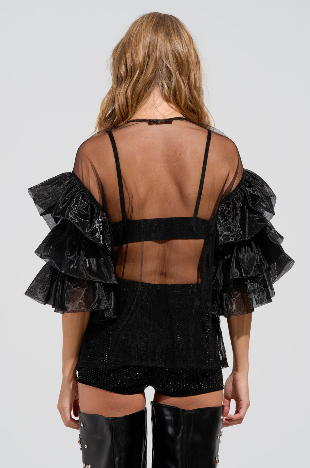 A person with long hair is seen from behind wearing the LOVE HARD TOP, a black sheer mesh knit top with layered ruffle detailing on the sleeves, paired with a sleek black bralette. They complete the look with black shorts and thigh-high black leather boots, achieving an effortlessly edgy style.