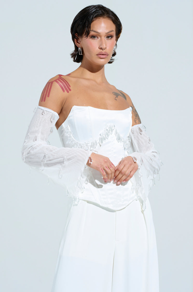 In a light blue backdrop, someone confidently showcases the KRISTENS DREAM SLEEVES—an off-shoulder white dress featuring sheer mesh knit sleeves and a fitted bodice. They flaunt short styled hair with visible arm tattoos.