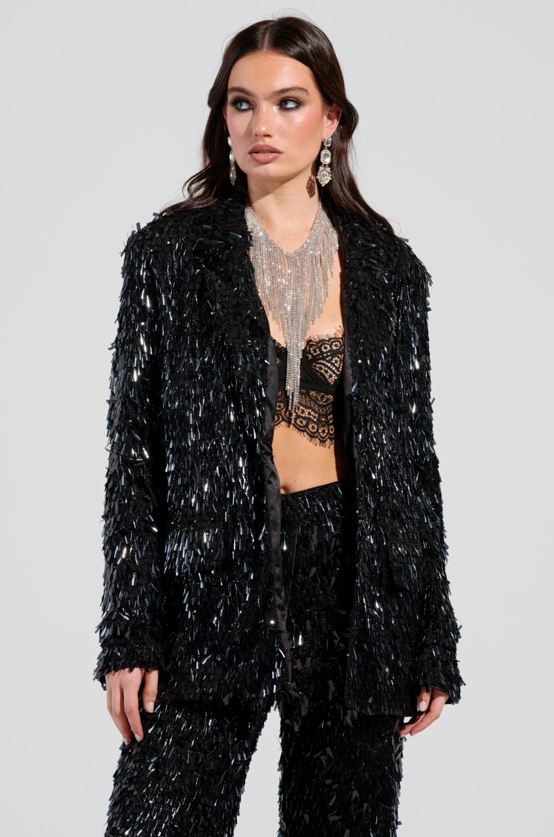 The woman wears a shimmering black outfit with the BLINGED OUT BLAZER by AKIRA Label and a lace top, standing against a plain background. Her intricate silver necklace and long earrings enhance her style, while her hair falls in loose waves, amplifying the ensemble's allure. 0