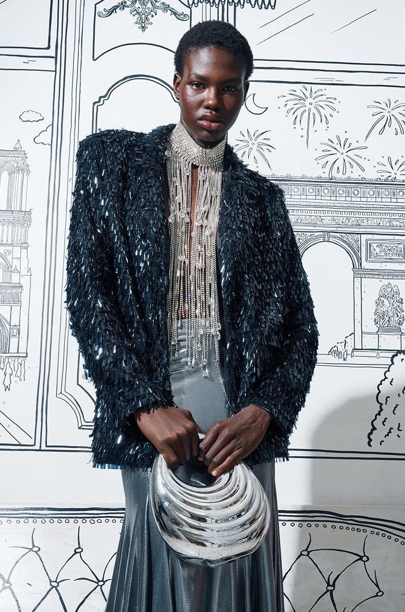 A person dressed in the BLINGED OUT BLAZER, combined with a silver skirt and an intricately beaded necklace, stands against line art architectural illustrations. They carry a shiny, reflective handbag that perfectly complements their dazzling outfit. 1