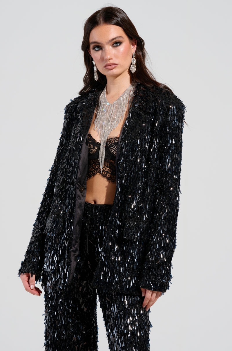 A woman poses in the "BLINGED OUT BLAZER" from the AKIRA Label, highlighting its textured, reflective sequin design. She pairs it with a black lace top and a statement necklace featuring dangling chains. Her dark hair is styled down, complemented by large earrings. 2