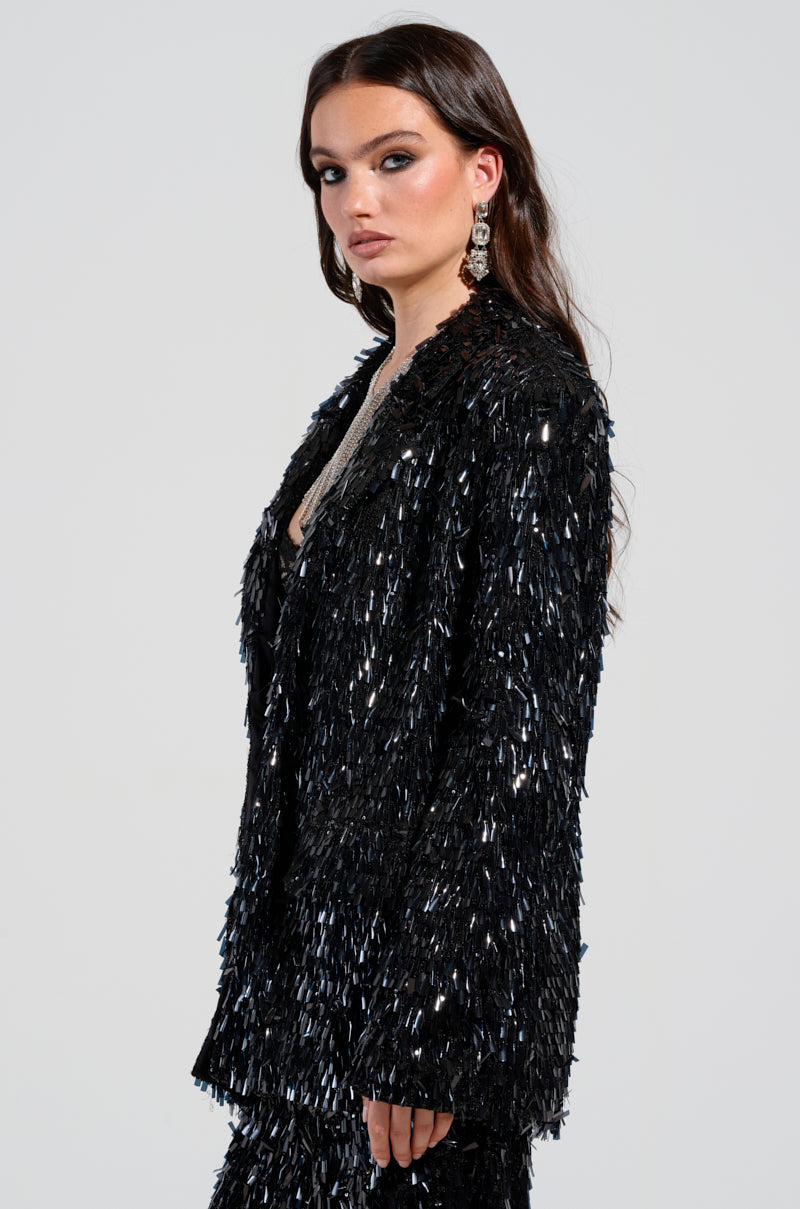 A person with long hair wearing the black, textured BLINGED OUT BLAZER from the AKIRA Label stands against a plain background. They are turned slightly to the side, highlighting their dangling earrings. 3