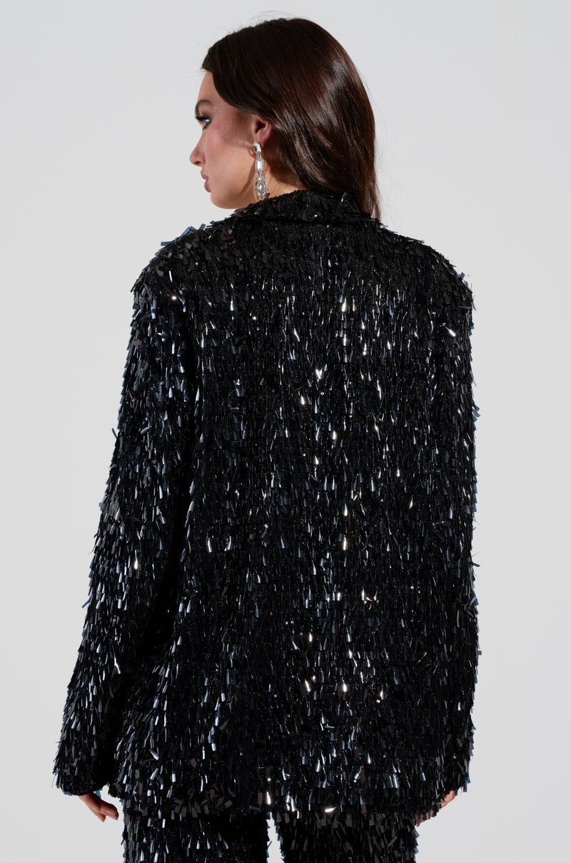 A woman with long dark hair is wearing the BLINGED OUT BLAZER from AKIRA Label, adorned with shimmering fringes. She is turned slightly to the side, highlighting the sparkly material. Additionally, she accessorizes with glittering drop earrings. 4