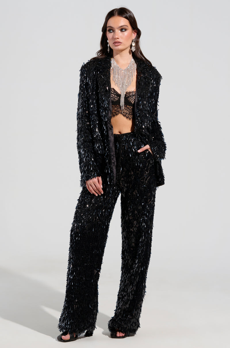 In an AKIRA Label textured suit, a woman stands confidently, showcasing her ensemble that includes a dramatic silver fringe necklace paired with a black lace bralette. Her look is enhanced by the BLINGED OUT BLAZER, while her long dark hair falls loosely around her as she poses against a plain, light background. 5