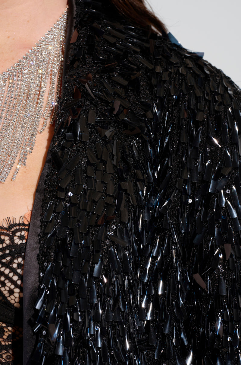 Close-up of a person wearing the BLINGED OUT BLAZER from AKIRA Label, shimmering in black with a glittering silver necklace of multiple strands. The blazer showcases intricate lace detailing visible underneath. 8