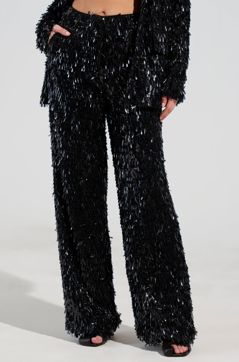 A person wearing the BLINGED OUT TROUSER, a shimmering black fringed pantsuit with sequin pants, stands against a plain background. The suit is adorned with long, textured fringes, and the person complements the look with black open-toed shoes.