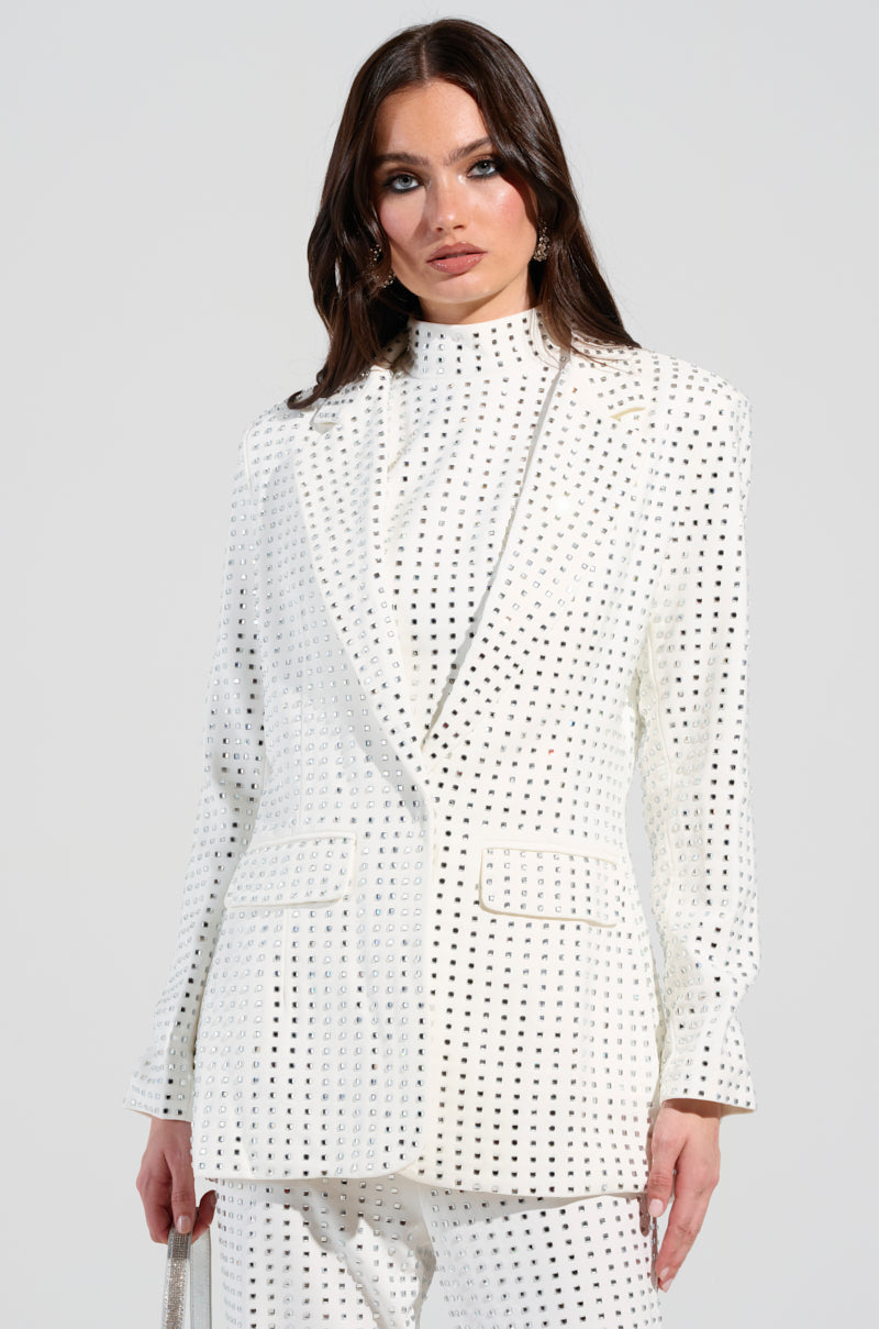 Dressed in an ICED OUT BLAZER, the individual pairs a white, checkered suit adorned with tiny black squares against a neutral backdrop. The semi-stretchy polyblend fabric beautifully accentuates their silhouette. Their long, dark hair flows gracefully as they hold a handbag at their side, while the silver rhinestones subtly elevate the ensemble.