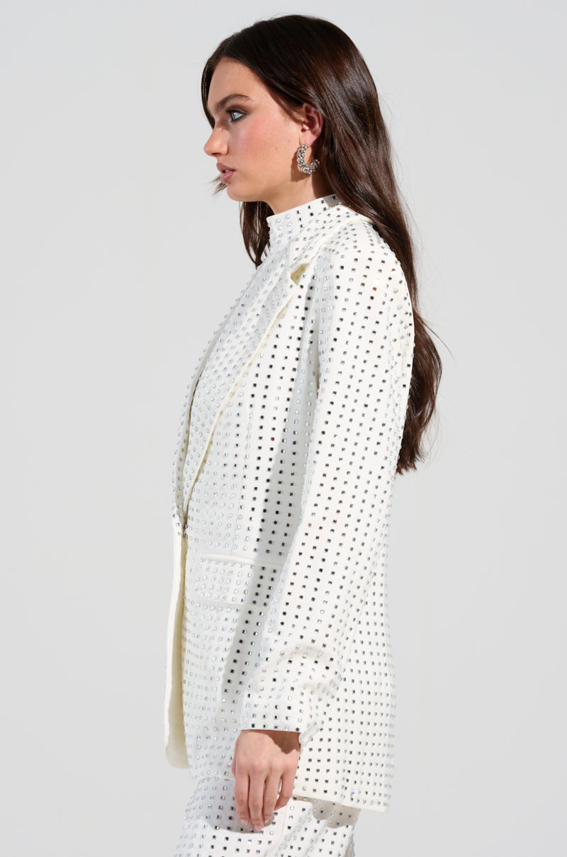 A person with long brown hair is wearing the ICED OUT BLAZER embellished with silver rhinestones. As they look to the left with a neutral expression, their hoop earrings add to their stylish appearance. The blazer, made from a semi-stretchy polyblend material, is striking against the plain, light-colored background.