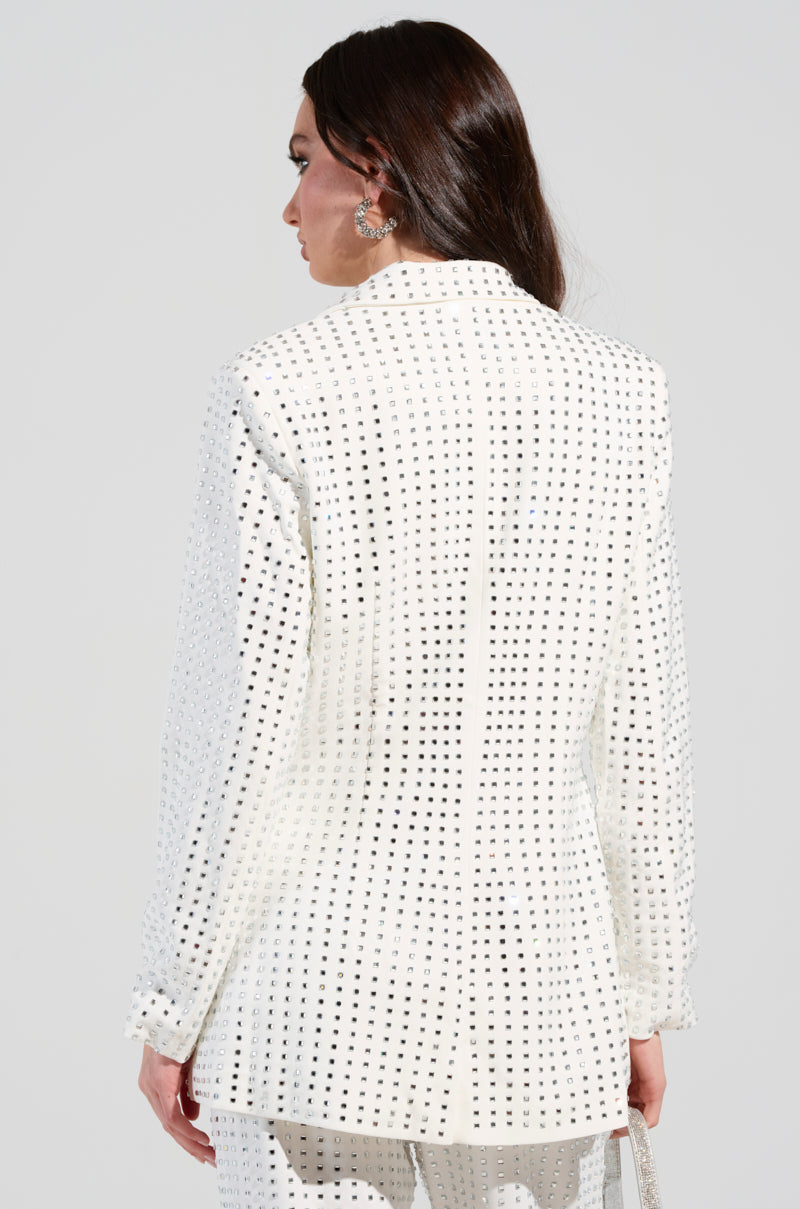 A person with long dark hair stands with their back facing the viewer, wearing the ICED OUT BLAZER - a chic white suit featuring black square patterns. They glance over their shoulder to highlight the suit's semi-stretchy polyblend back design, elegantly adorned with silver rhinestones. The backdrop is a simple light gray.
