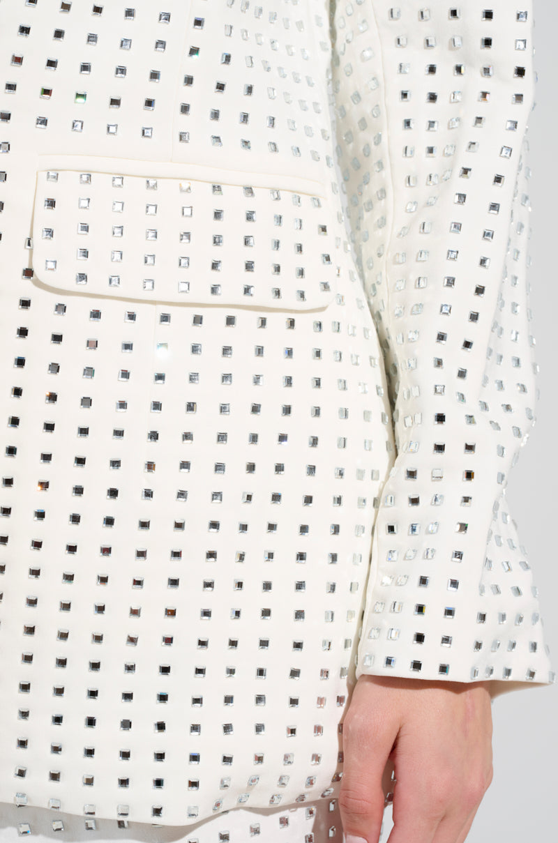 Close-up of a person wearing the ICED OUT BLAZER, a white jacket adorned with square silver studs and shimmering rhinestones. Made from a semi-stretchy polyblend, it boasts a visible pocket and sleeve, with the person's hand gently resting near the pocket against a plain, light-colored background.