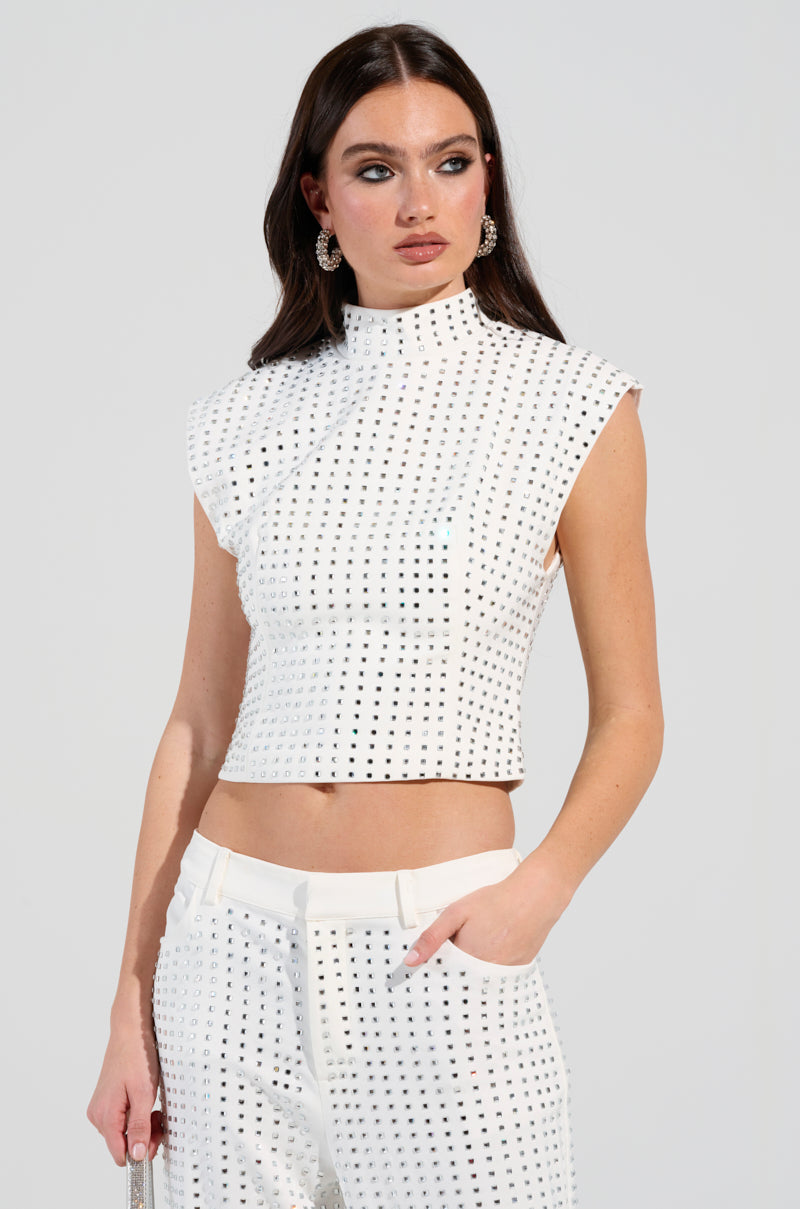 A woman wearing the ICED OUT TOP, embellished with rhinestones, pairs it with matching pants and poses confidently with one hand in her pocket. She accessorizes with large hoop earrings against a plain background.
