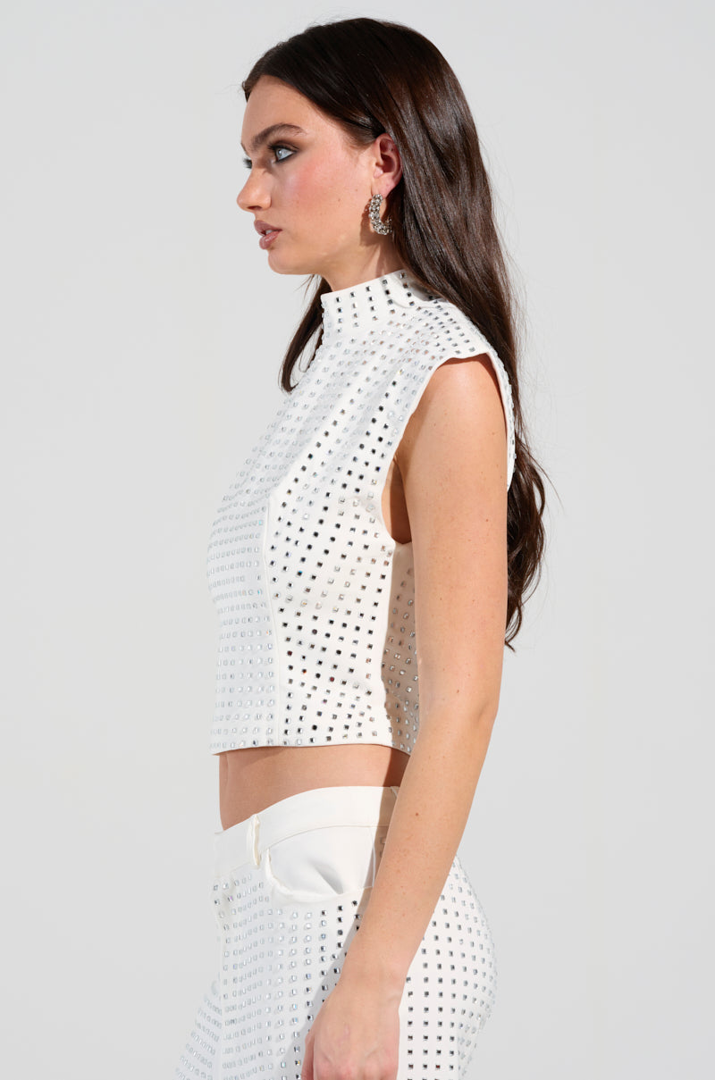 A woman with long dark hair is wearing a sleeveless white ICED OUT TOP embellished with rhinestones, paired with matching high-waisted pants. She is facing left against a plain background, and the metallic embellishments add a touch of sparkle.