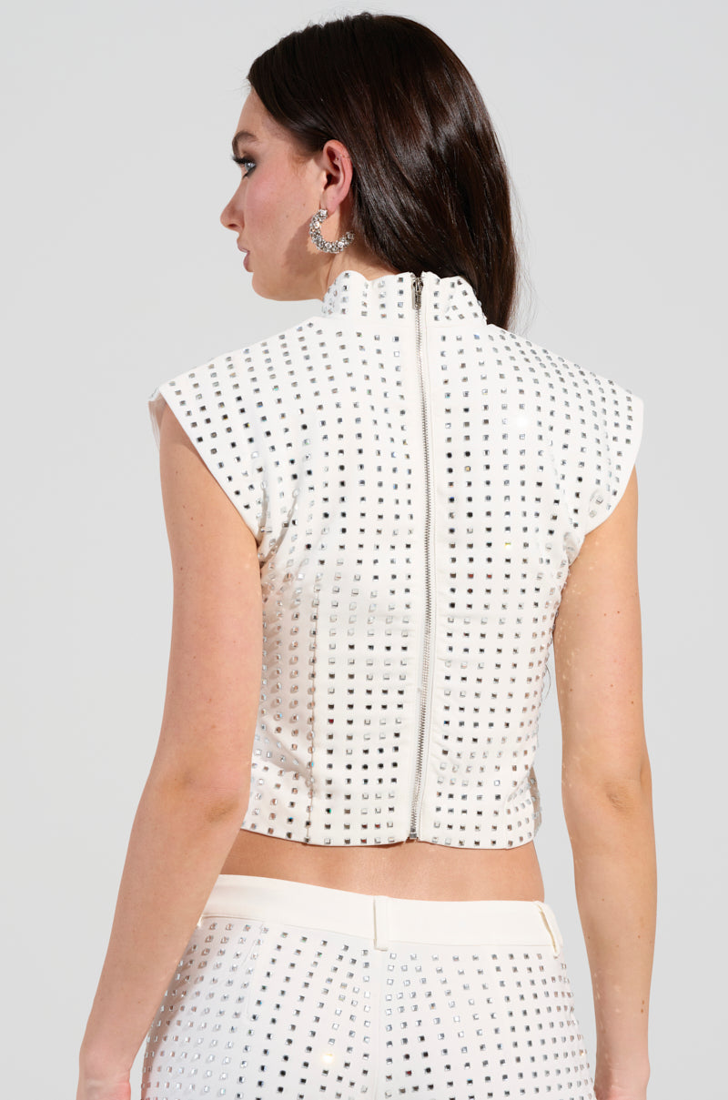 A woman with long brown hair is dressed in an ICED OUT TOP paired with matching pants, both adorned with small metallic studs. The ensemble showcases a high collar accentuated by shimmering rhinestones, and she is positioned slightly turned away from the camera.