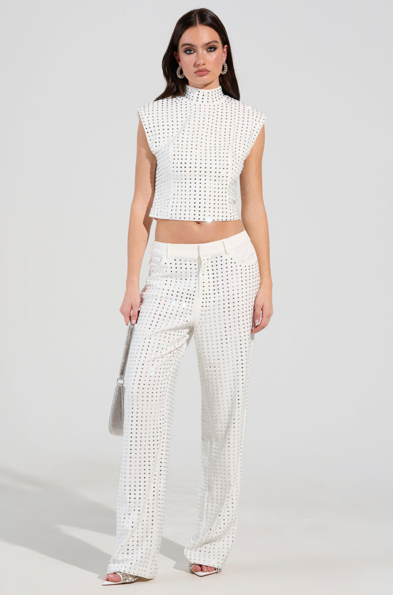 A woman stands wearing the ICED OUT TOP embellished with rhinestones, paired with matching wide-leg pants. She carries a silver purse, her long dark hair cascading down her shoulders. The plain, light-colored background accentuates her chic ensemble.