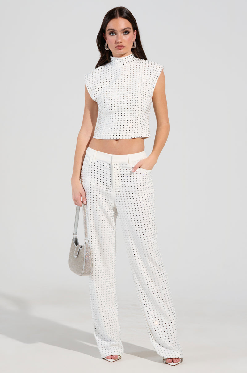 A person in a sleeveless white top and high-rise pants adorned with black dots strikes a pose. They carry a silver handbag inspired by the ICED OUT TROUSER and sport rhinestone-embellished silver sandals, all set against a simple light background.