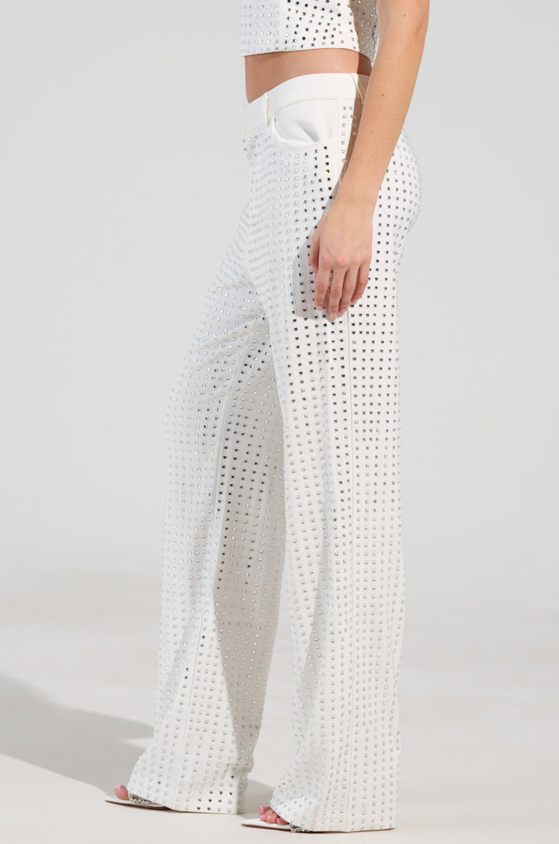 A person wearing an all-white outfit with the ICED OUT TROUSER featuring a matching crop top. The high-rise trousers are embellished with rhinestones in a grid pattern, creating a shimmering effect against the plain and minimalistic background.
