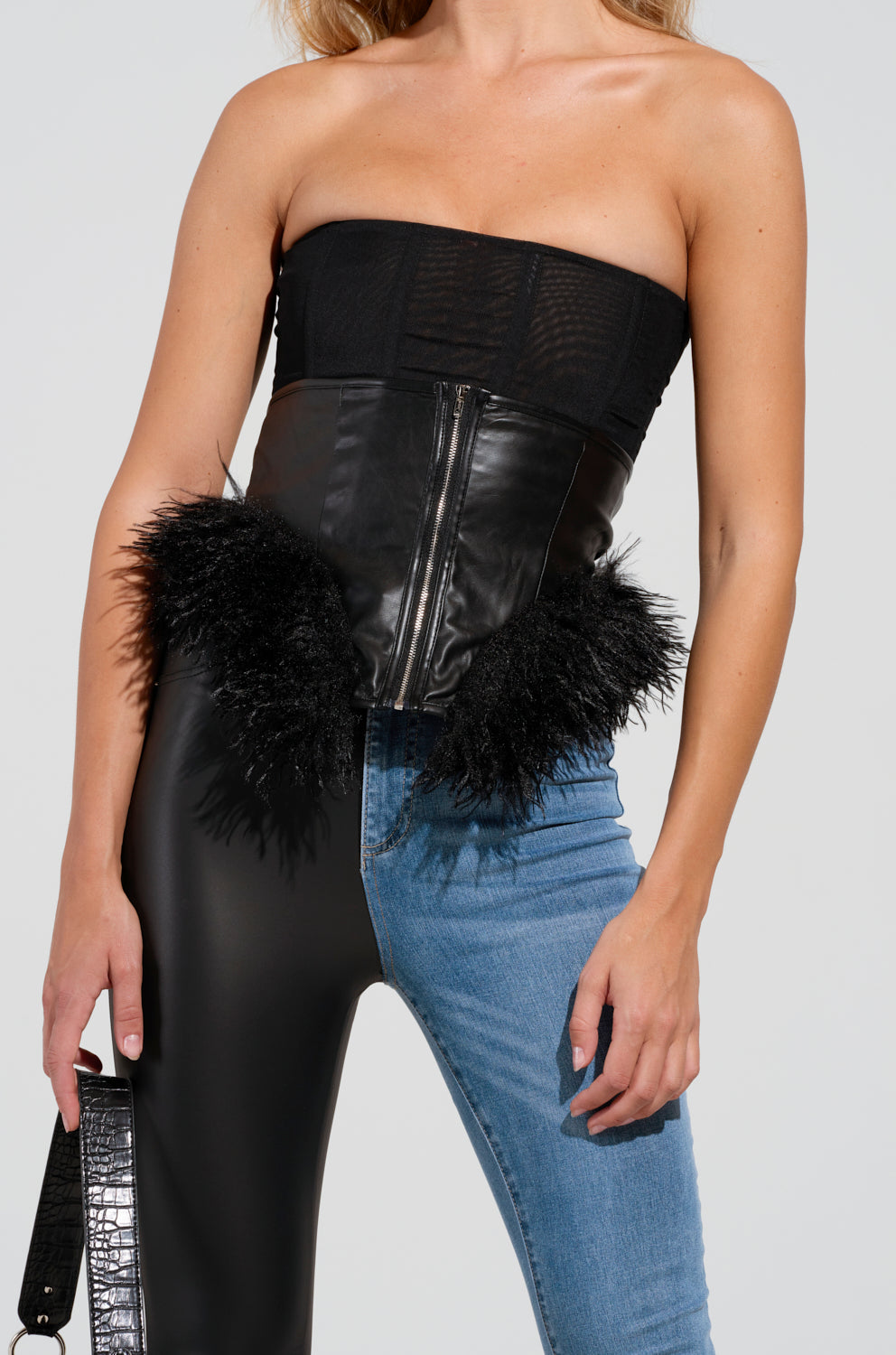 A woman in the XENON faux fur trim black leather top with feather detailing pairs it flawlessly with asymmetrical pants—half shiny black, half denim. She holds a textured black bag, all set against a plain gray background.