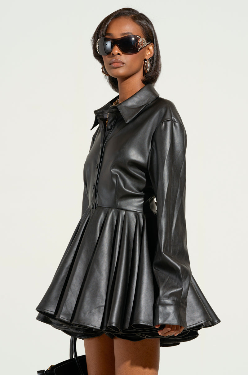 A person confidently poses against a plain light background, wearing THE IT GIRL MINI DRESS IN BLACK PU with a flared skirt and large sunglasses. Statement earrings complement the stylish ensemble, adding a touch of elegance.