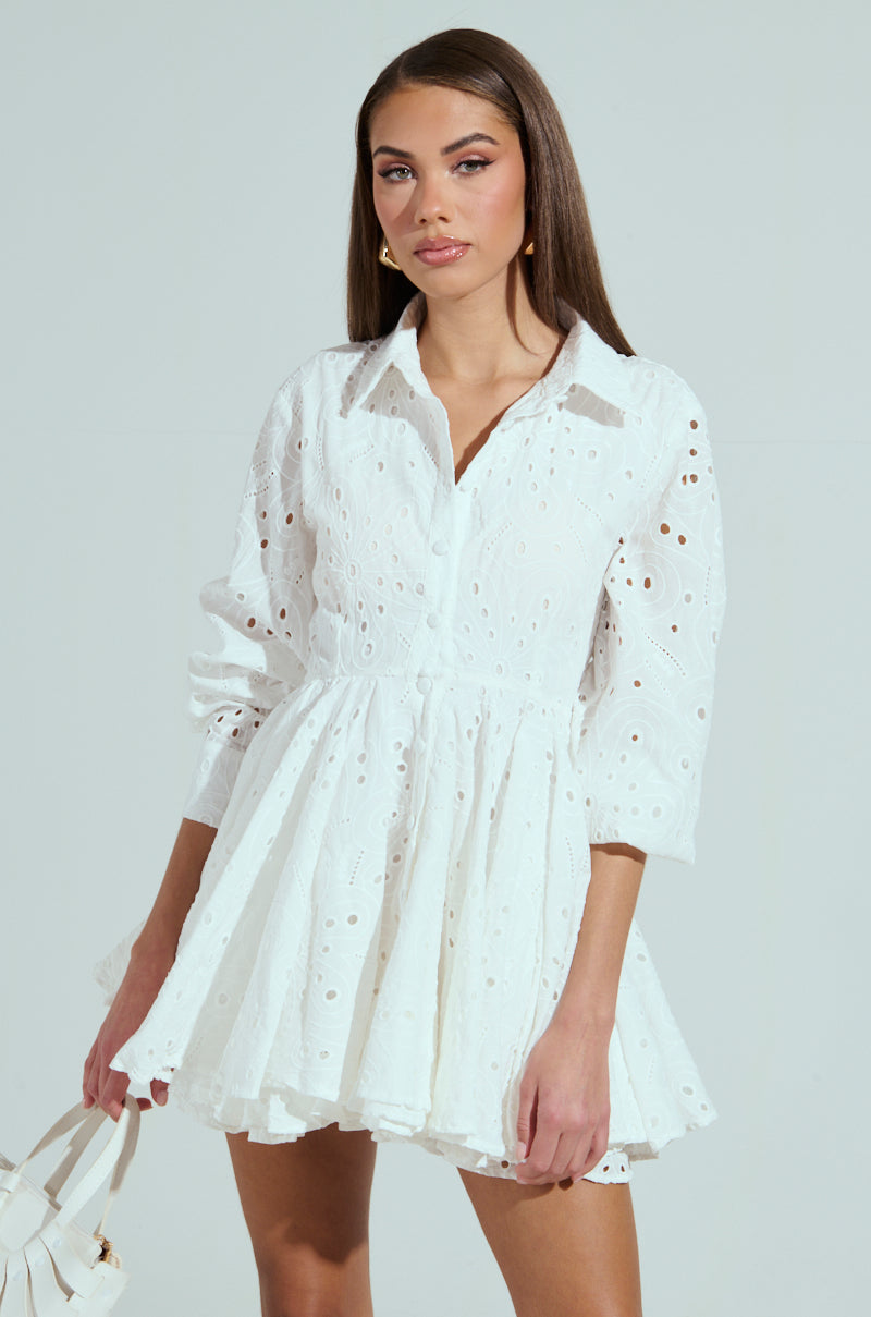 A woman stands against a light background wearing THE IT GIRL MINI DRESS IN EYELET, a white lace dress with long sleeves and a flared skirt. She carries a white handbag, has long straight brown hair, and maintains a neutral expression.