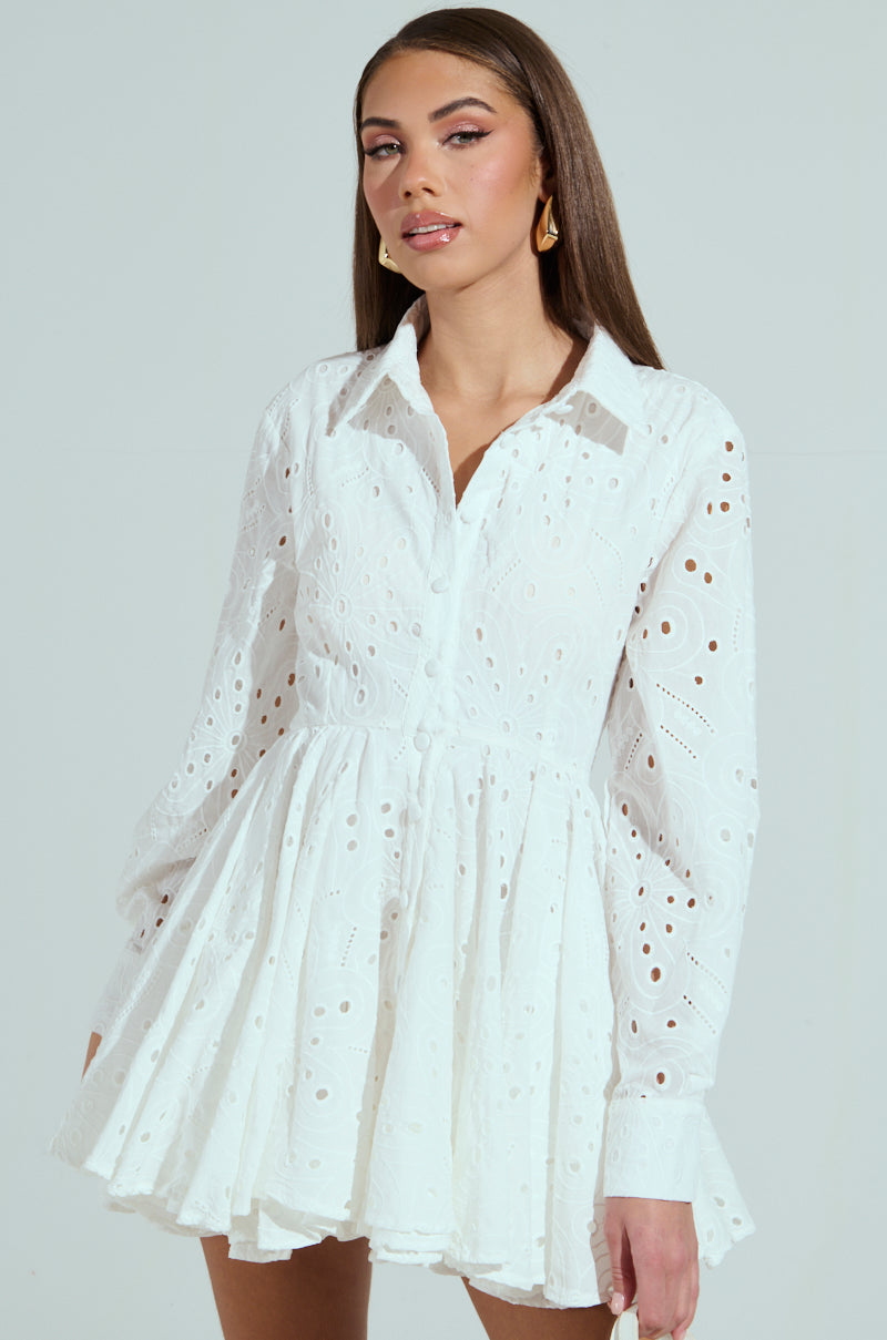 A woman confidently poses in THE IT GIRL MINI DRESS IN EYELET, a white long-sleeve mini with a flared skirt. Her long, straight hair complements her gold hoop earrings against a simple light gray backdrop.