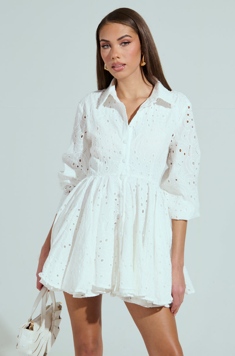A person with long brown hair is wearing "The It Girl Mini Dress in Eyelet" and holding a white handbag, standing against a light gray background, embodying effortless style.