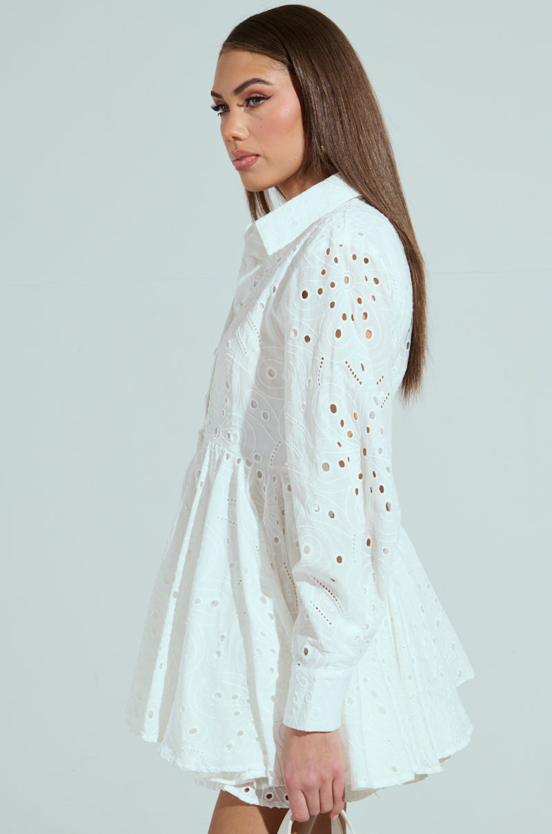 A woman with long brown hair stands sideways in THE IT GIRL MINI DRESS IN EYELET, a white, long-sleeved cotton frock featuring eyelet patterns, intricate embroidery, and a flared skirt. The background is a soft pastel shade.