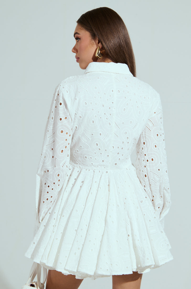 A woman with long brown hair dons THE IT GIRL MINI DRESS IN EYELET, a white, intricately patterned cotton frock with long sleeves and a flared skirt. She faces away, holding a small white bag against a light-colored backdrop.