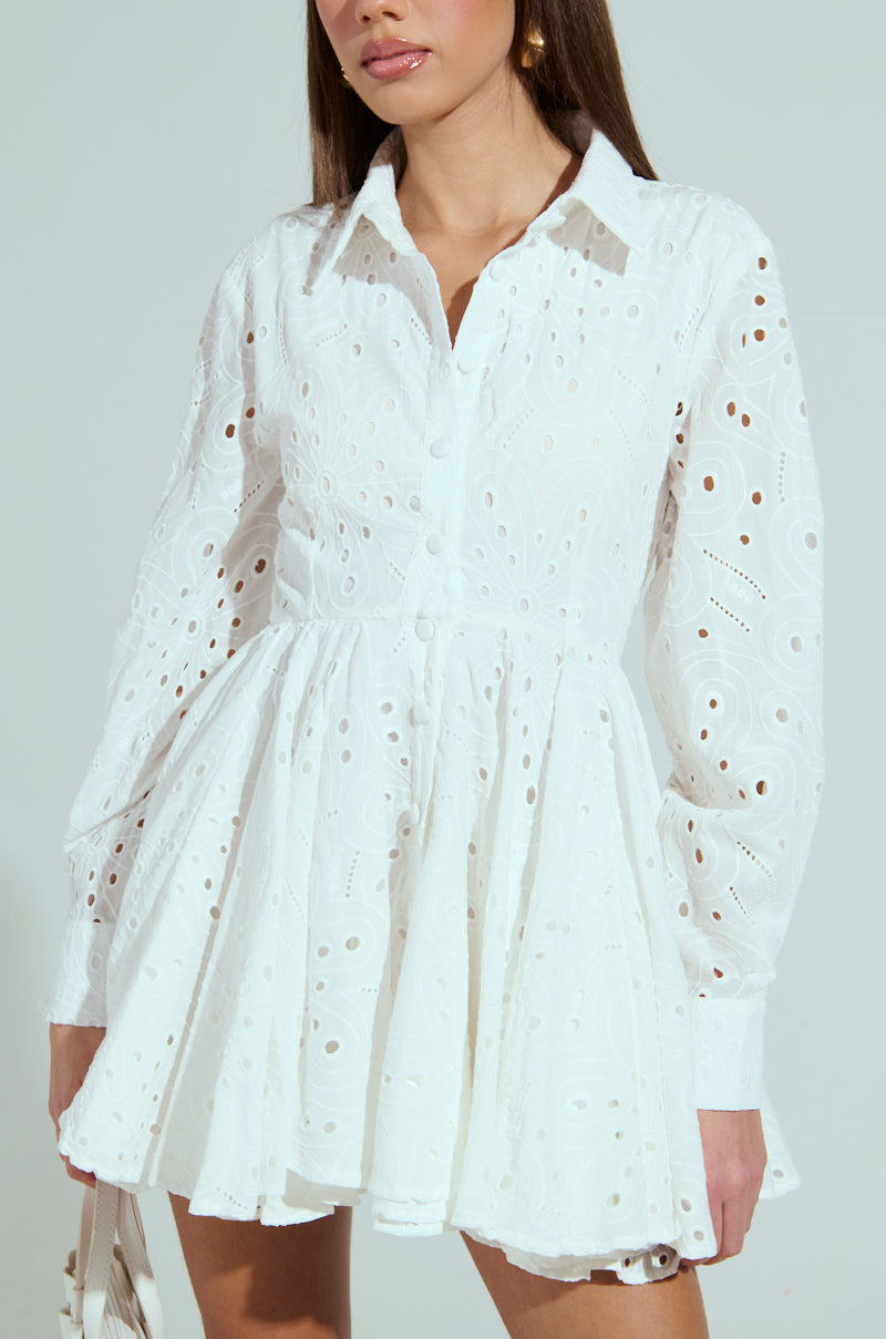A woman in the white, long-sleeved "THE IT GIRL MINI DRESS IN EYELET" with flared skirt and intricate embroidery holds a small handbag. The dress features front buttons as her hair cascades against a neutral gray backdrop, epitomizing the It Girl style.