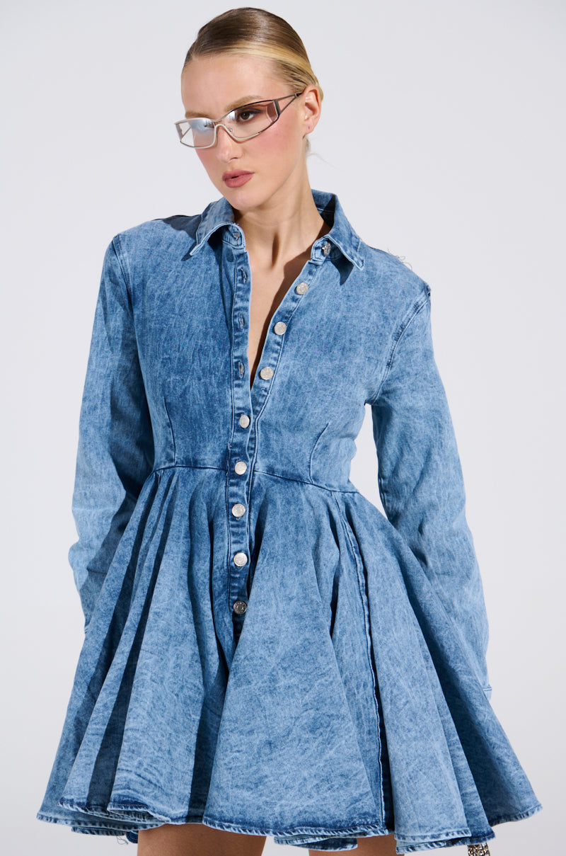 A person with sleek glasses models the AKIRA Label's THE IT GIRL MINI DRESS IN DENIM, featuring a blue button-down design and a flared skater skirt. Their slicked-back hair and the plain white background highlight this modern, stylish outfit.
