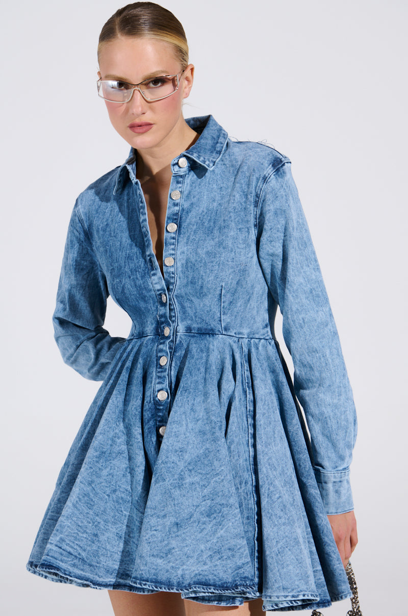 Someone confidently poses against a plain background in THE IT GIRL MINI DRESS IN DENIM from the AKIRA Label, with sleek hair pulled back and accessorized with stylish sunglasses.