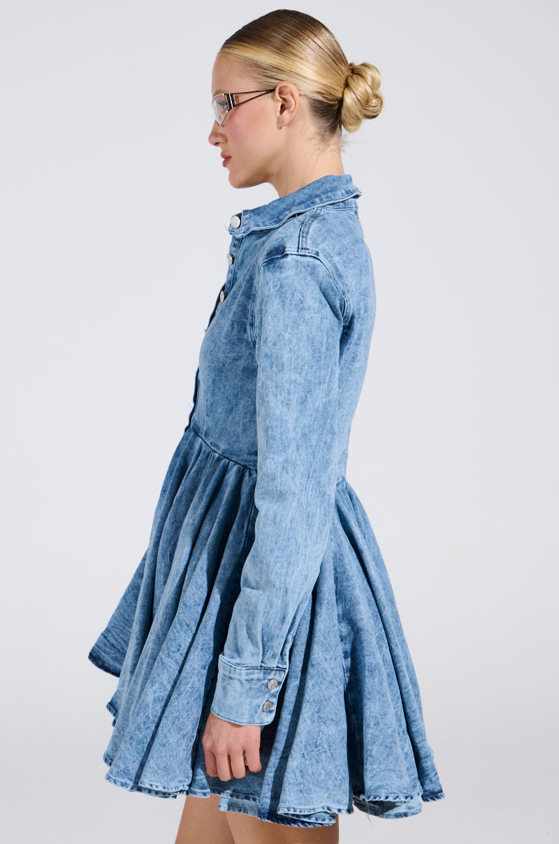 A person with glasses and a bun is wearing THE IT GIRL MINI DRESS IN DENIM, featuring a flared skirt. They are standing sideways against a plain, light background.