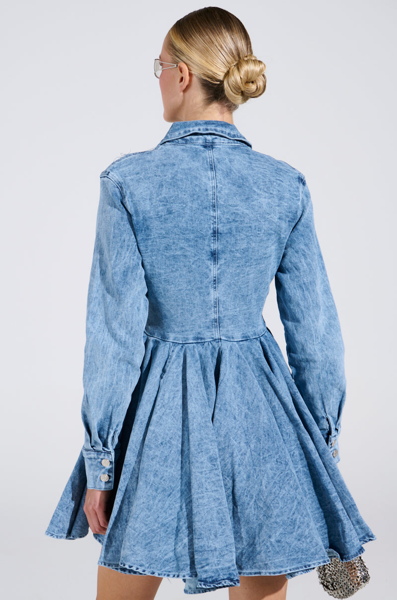 A person with blonde hair in a bun is wearing the THE IT GIRL MINI DRESS IN DENIM, featuring a flared skater skirt. They face away from the camera against a plain light background.