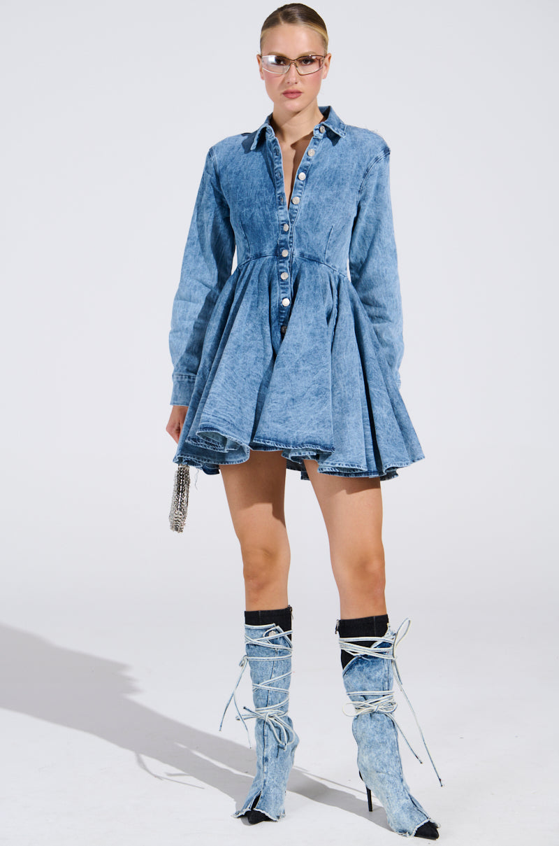 A person poses in "THE IT GIRL MINI DRESS IN DENIM," featuring a flared skater skirt and high denim boots with white laces. Wearing sunglasses and carrying a small purse, they stand against a plain white background.