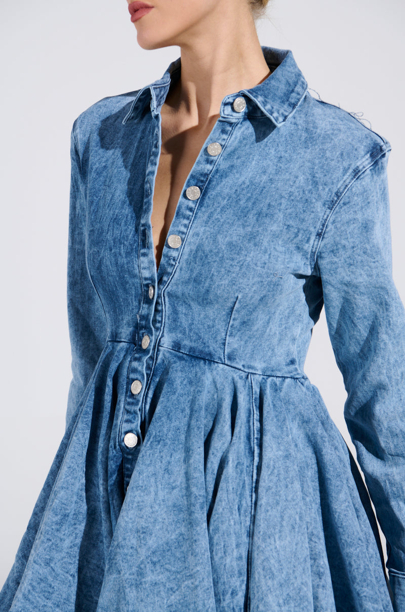 Wearing the IT Girl Mini Dress in Denim, a stylish button-up with pleated skater skirt. Silver buttons and a smart collar highlight the fitted waist, all on a sleek white background.