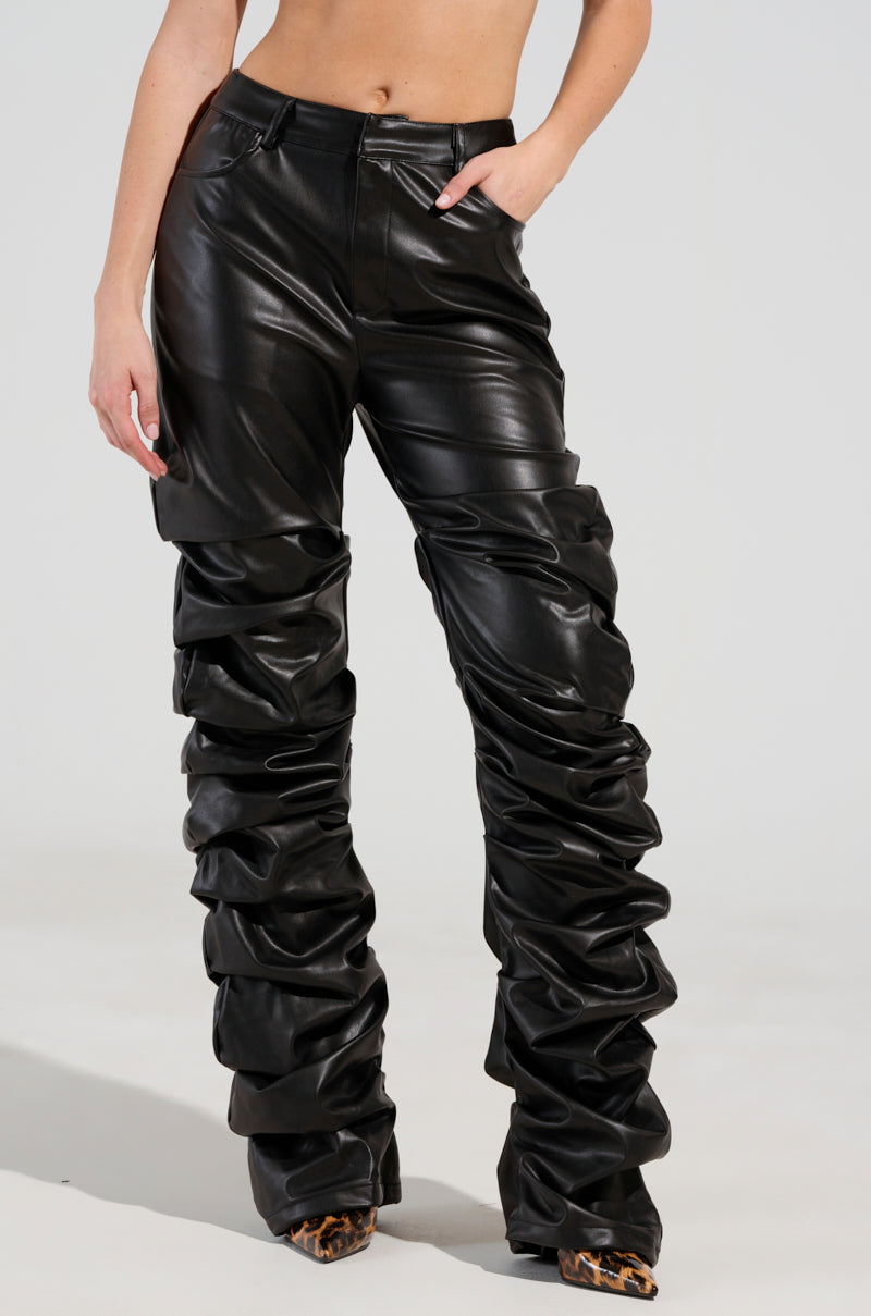 A person wearing the DOUBLE TROUBLE TROUSER in black faux leather, designed with an exaggerated, gathered pattern down the legs for a sleek skinny silhouette, stands with one hand in a pocket. The ensemble is completed with pointed leopard-print shoes.