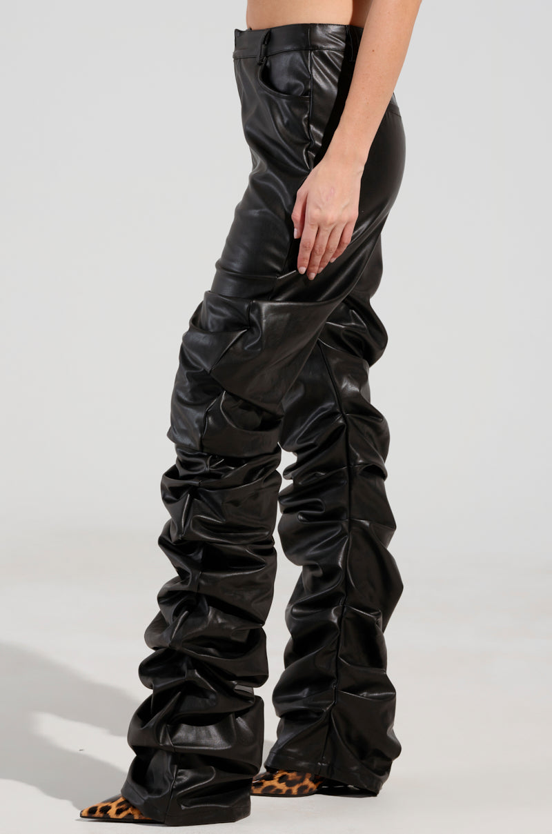 A person models the DOUBLE TROUBLE TROUSER, featuring high rise, black ruched faux leather material with flared legs, paired with animal print shoes against a plain background.