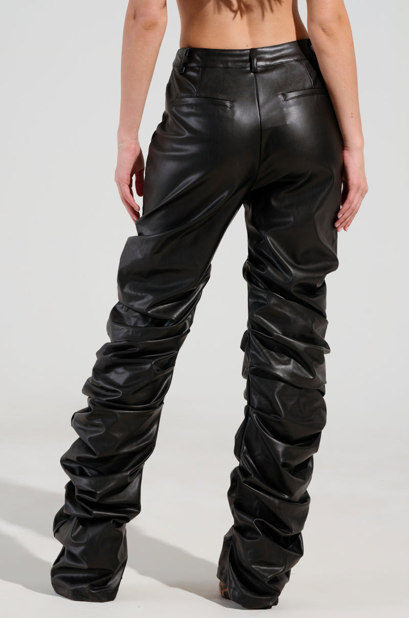 Rear view of a person wearing the DOUBLE TROUBLE TROUSER, featuring high-rise faux leather with a glossy finish. The snug fit transitions into a loose, gathered effect along the legs, creating a distinctive skinny leg silhouette. The person's arms are bare and positioned at their sides.