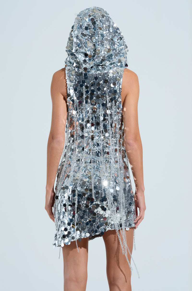 A person stands with their back to the camera, wearing the WON OF ONE SEQUIN CHAIN MINI DRESS. This sleeveless outfit features fringe details and a hood, creating a striking silhouette against the plain, light-colored background.