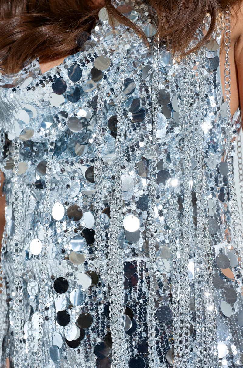 A close-up features the WON OF ONE SEQUIN CHAIN MINI DRESS sparkling with silver embellishments. The stretchy mesh fabric shimmers, accentuating intricate details, with strands of the person's brown hair visible at the top.