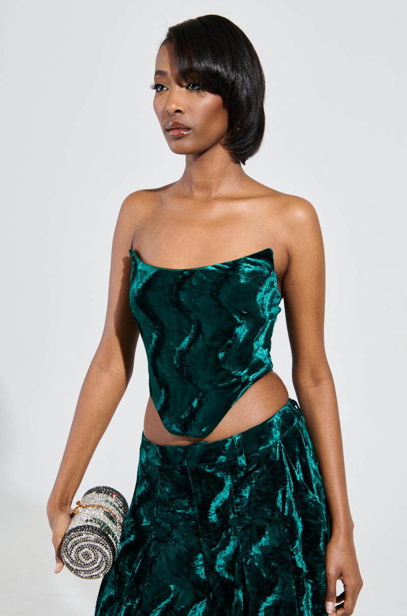 A person wearing the "GREEN WITH ENVY VELVET CORSET TOP" and a matching skirt poses with a small, beaded cylindrical clutch in their left hand. They have shoulder-length hair and a confident expression. 0
